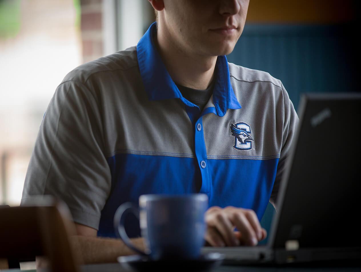 A man in a Creighton University polo typing on a laptop